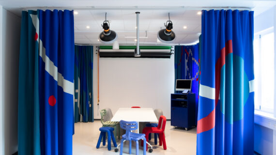 Photo de la salle d'innovation pédagogique située dans le bâtiment Pulse à la HES-SO Genève. La salle est installée en configuration atelier de travail pour un groupe de 6 personnes avec une grande table et des chaises autour. On aperçoit 3 écrans de projection non-déroulés au fond de la pièce. Tout autour de la pièce, un grand rideau bleu à motif entoure l'ensemble. Un ordinateur disposé sur le côté permet de piloter le matériel.